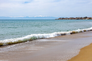 A swirling wave curves along the shoreline with wet sand and foam, backed by snow-capped mountains across Antalya Bay and the historic old town cape. Low-season tranquility in Sorgun, Antalya, Turkey.
