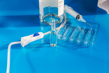 Roller clamp on IV drip next to medicine ampoule, syringe, and bottle with saline solution (a mixture of water and sodium chloride) against medication instruction and blue background, close-up.