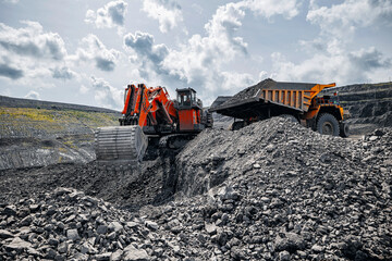 Red excavator loads black coal into back of yellow heavy mining dump truck. Open pit mine process, industry landscape.
