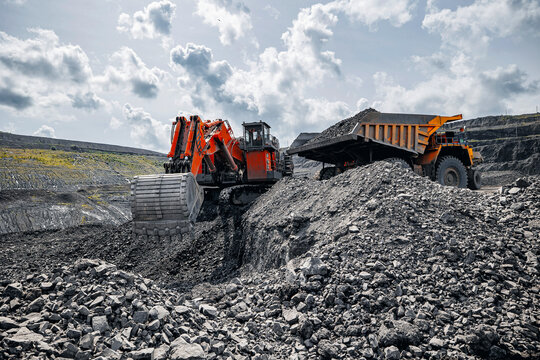 Red excavator loads black coal into back of yellow heavy mining dump truck. Open pit mine process, industry landscape.