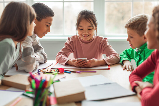 Diverse group of school kids sit at a desk in their classroom, having fun while using a smartphone. They are engaged in an educational app and sharing laughter during their break.