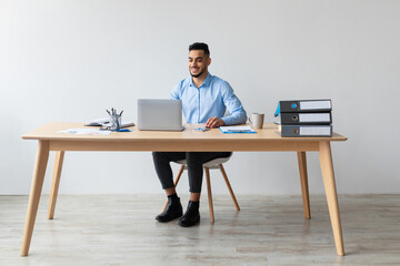 Smiling Arab manager engages with his laptop in a contemporary office. He enjoys watching a webinar while seated at his desk.