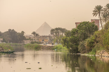 View of Great Pyramid of Giza through haze across canal water and green vegetation