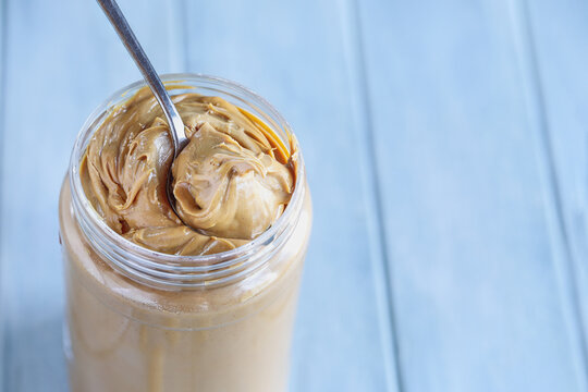 Peanut butter being scooped up with a spoon from a plastic jar container over a blue table. Selective focus with blurred background. 