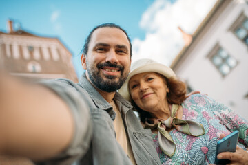 A pensioner and her adult son on a guided tour of an ancient castle. They smile happily and take selfies on their phones. An elderly mother and son travel together, visiting historical sites