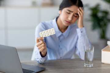 Young Indian woman experiences severe pain and discomfort at her workplace. She is seated at a desk with a laptop and holds a strip of pills, reflecting her struggle with migraine and illness.
