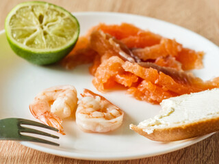 A fork near a big shrimp, and the plate holds sliced salmon, cream cheese on bread, and a lime