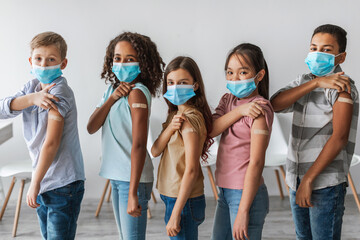 Obraz premium Children from different backgrounds display their arms with medical bandages after getting the Covid-19 vaccine. They are indoors, wearing masks and standing against a gray wall.