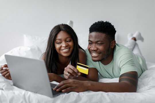 A smiling couple is lying on a cozy bed while they look at a laptop. One partner holds a credit card, suggesting they are shopping online. The setting is bright and inviting.