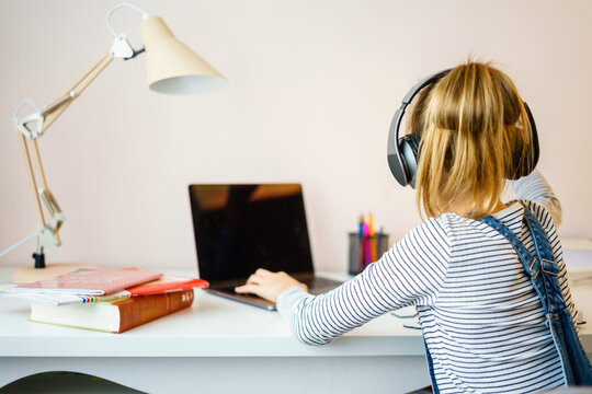Portrait of teenage girl learning online with headphones and laptop taking notes in a notebook sitting at her desk at home doing homework
