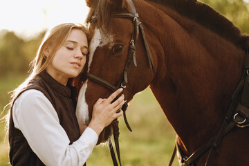 Obraz premium Young beautiful female jockey strokes and hugs the horse's head and prepares for the competition. Jumping training in the meadow in summer evening.