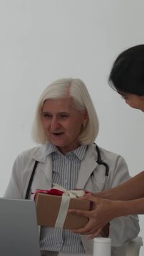 Vertical video diverse medical team surprises senior female physician in her office with gifts, hugs and smiles to celebrate a major career milestone, gratitude and warm congratulations