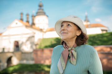 Пожилая довольная женщина гуляет на экскурсии в парке возле старинного замка. Пенсионерка рассматривает историческую архитектуру.
