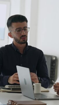 Vertical video middle eastern male doctor with glasses speaking confidently at a laptop during a collaborative medical team meeting in hospital office