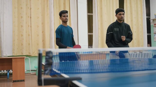 Two young athletes during indoor table tennis practice session, standing near net with paddles in hand, preparing to return ball in competitive and concentrated atmosphere under bright hall lighting