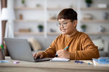 Asian teen boy sits at a table with stationery in front of a modern laptop, doing homework and studying at home during COVID-19, focused on his online lesson and taking notes.
