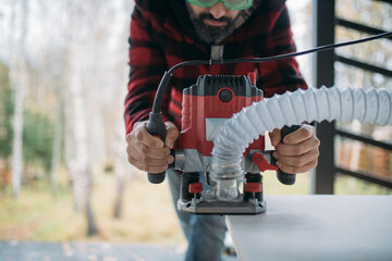 A young man is working with a milling cutter on the veranda of a house. The carpenter chamfers the plywood using a milling machine.