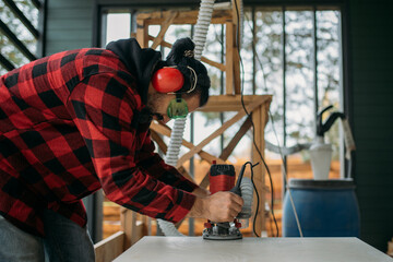 A young man is working with a milling cutter on the veranda of a house. The carpenter chamfers the plywood using a milling machine.