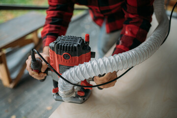 A young man is working with a milling cutter on the veranda of a house. Close-up of hands holding the machine. The carpenter is chamfering plywood using the milling machine.
