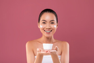 Young korean woman displays her top facial product with a cheerful smile. She uses hydrating cream...