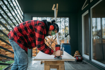 A young man is working with a milling cutter on the veranda of a house. The carpenter chamfers the plywood using a milling machine.