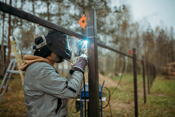 A young man is working as a gas welder on the street. A man wearing a protective mask is welding iron rungs to pillars. The worker, holding a welding machine, sparks fly around him.