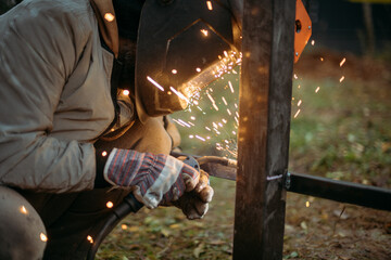 A young man is working as a gas welder on the street. A man wearing a protective mask is welding iron rungs to pillars. The worker, holding a welding machine, sparks fly around him.