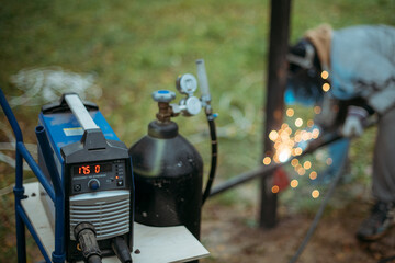 A young man is working as a gas welder on the street. A man wearing a protective mask is welding iron rungs to pillars. The worker, holding a welding machine, sparks fly around him.