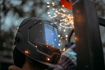 A young man is working as a gas welder on the street. A man wearing a protective mask is welding iron rungs to pillars. The worker, holding a welding machine, sparks fly around him.
