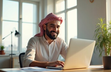 Arab man wears keffiyeh smiles while typing on laptop. He works remotely from home office, sitting at desk with plant and lamp nearby. Successful young freelancer enjoys his modern workspace.