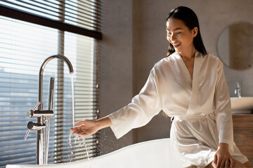 A positive and attractive young woman in a silky bathrobe sits comfortably in a modern bathtub. She is smiling as she checks the water temperature in a bright bathroom.