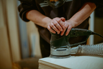 A young woman grinds plywood in a house under construction. Close-up of a woman's hands holding a grinder. A woman works with a manual electric grinder