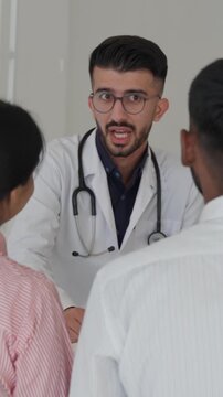 Vertical video middle eastern doctor in professional attire, including a white lab coat and stethoscope, discussing a diagnosis and giving medical guidance to a couple in a hospital setting