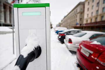 Electric vehicle charging station covered in snow during winter with visible cable and connector, sustainable mobility in harsh weather conditions. Parking lot for electric vehicles