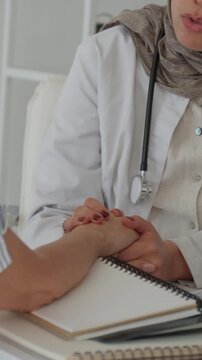 Vertical video muslim woman doctor in hijab and lab coat offers comforting hand and empathetic support to patient during medical consultation