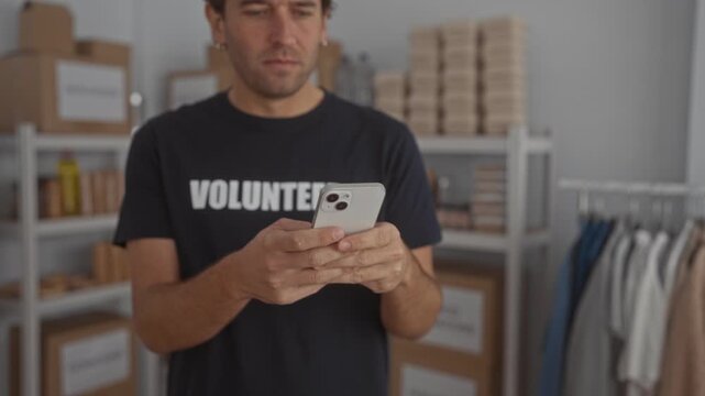 Hispanic volunteer man in black shirt using smartphone in organized room with shelves and clothes, symbolizing tech-savvy aid work in a structured environment.