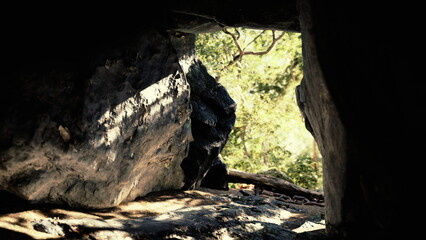 Soft light filters through a rocky opening, revealing a lush, green forest. Shadows dance on the ground as nature thrives outside this secluded space, creating a peaceful atmosphere.