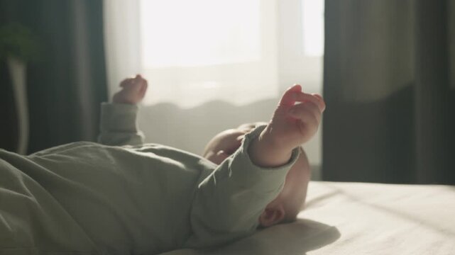 Three month old baby boy in bodysuit lies on a bed in morning sun lights. Newborn fingers in selective focus. Funny playful and energetic child waving hands slow motion