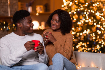 A young african american couple sits on a cozy sofa, smiling and sipping from red cups. They enjoy...