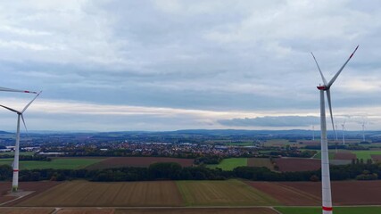Tall wind turbines dot the green fields, harnessing wind energy under a moody sky. A small village lies in the background, surrounded by vibrant autumn colors. - Powered by Adobe