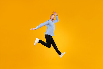 Fototapeta premium Mature man reacts with joy, jumping high in an orange studio. His expression shows excitement as he celebrates a triumph, dressed casually in a blue checkered shirt and black pants.