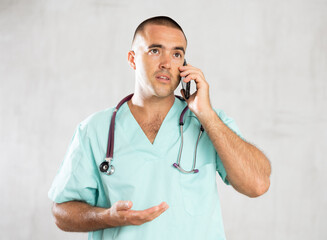 Focused doctor wearing white coat with phonendoscope on neck talking on phone in medical clinic office