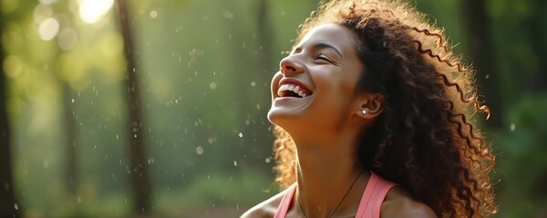 Fototapeta premium Young happy woman with curly hair laughs outdoors in a sunny park. She feels pure joy, freedom, and positive energy in nature. Light mist falls around her, enjoying fresh air and green trees.