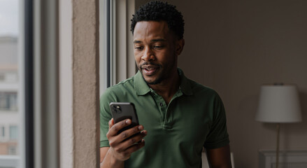 young man in green shirt smiling while reading message on smartphone by window