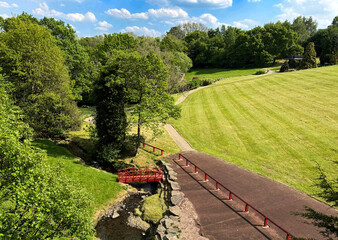 A lush green park cradles a bright red bridge spanning a quiet stream, while the open sky drifts with a few idle clouds near Thornton Road, Bradford, UK