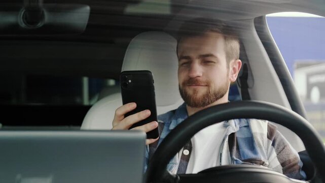 Smiling young man sitting in the car and looking at phone screen. Slow motion