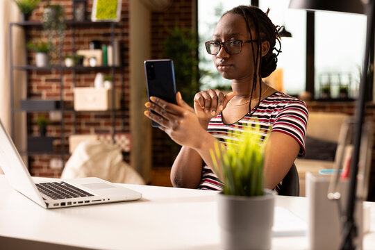 Self employed woman holds phone during remote team meeting, reviewing performance metrics in home office. Focused entrepreneur on video call via smartphone, discussing company data and operations.