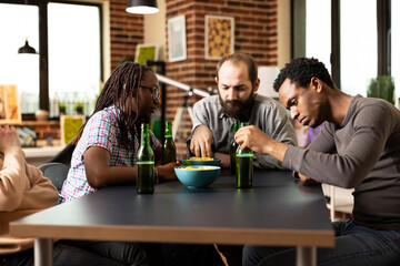 Young male and female adults sit around table with beer and chips, looking bored in cozy room. Group of friends relax together in brick walled room, sharing snacks and drinks, feeling unoccupied.