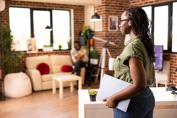 African american woman walking through brick wall room with laptop in hand, preparing to work from home. Black female freelancer standing with digital device as boyfriend sits in background with book.