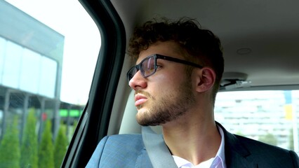 Calm young businessman in glasses sitting on back seat of taxi car while looking out the window. Business, trip, transport and people concept. Slow motion